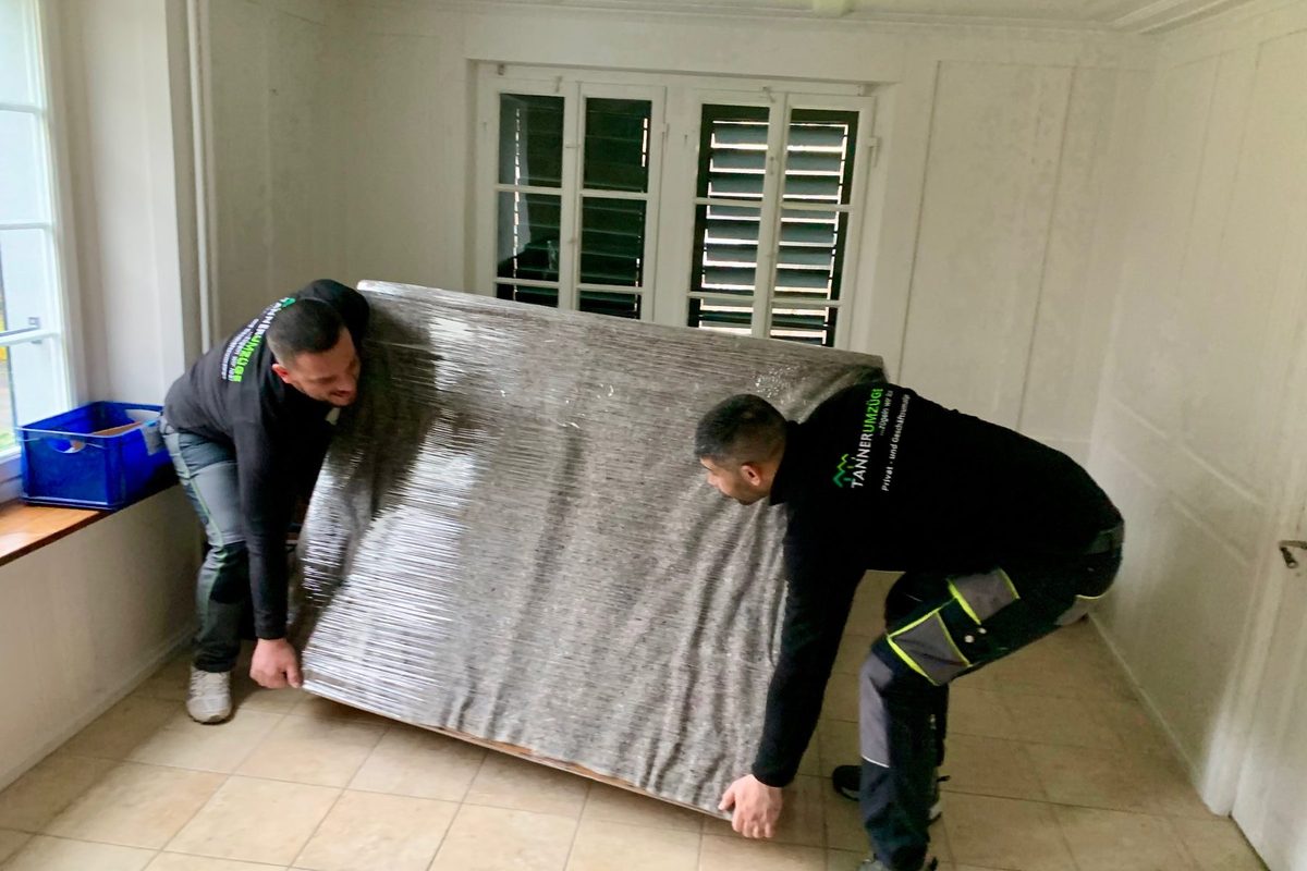 Zwei Mitarbeiter von Tanner Umzüge tragen eine in Folie eingepackte Matratze aus dem Zimmer